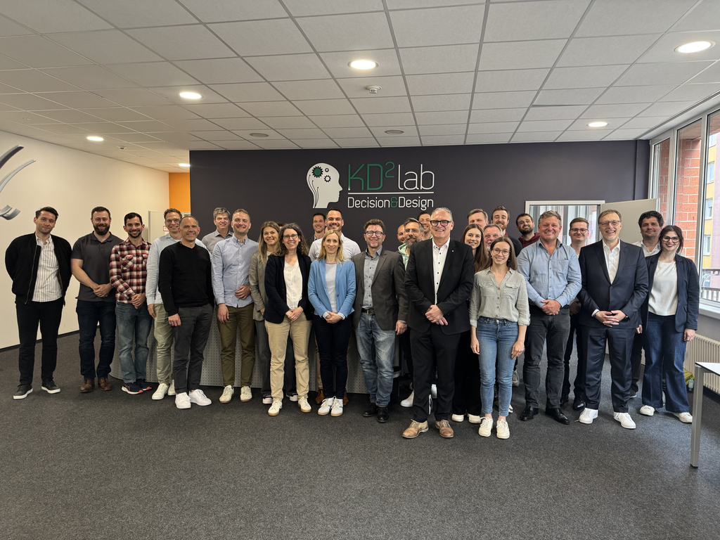 Gruppenfoto von Mitarbeitern im KD²Lab vor einer Wand mit Logo und Schriftzug "Decision Design".